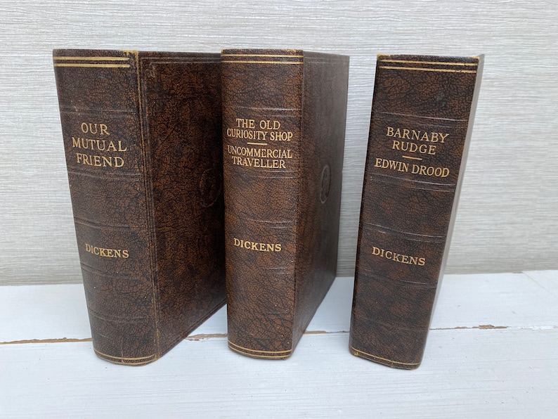 May include: Three antique books with brown leather covers. The titles "Our Mutual Friend", "The Old Curiosity Shop", and "Barnaby Rudge" are printed in gold. The author, Dickens, is also printed on the spines.