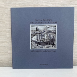 May include: A square, blue-gray book cover with the title "Rudyard Kipling's The Shipwright's Trade." The cover features a black and white illustration of a boat and a person. The text "with wood & linocuts by James Dodds" and "Jardine Press" are also visible.