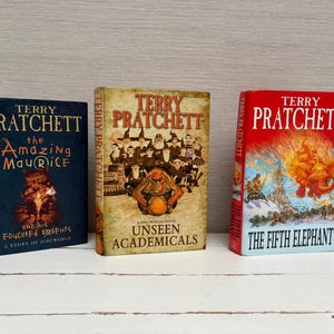 May include: Three hardcover books by Terry Pratchett, featuring colorful illustrations and titles: "The Amazing Maurice and his Educated Rodents", "Unseen Academicals", and "The Fifth Elephant".