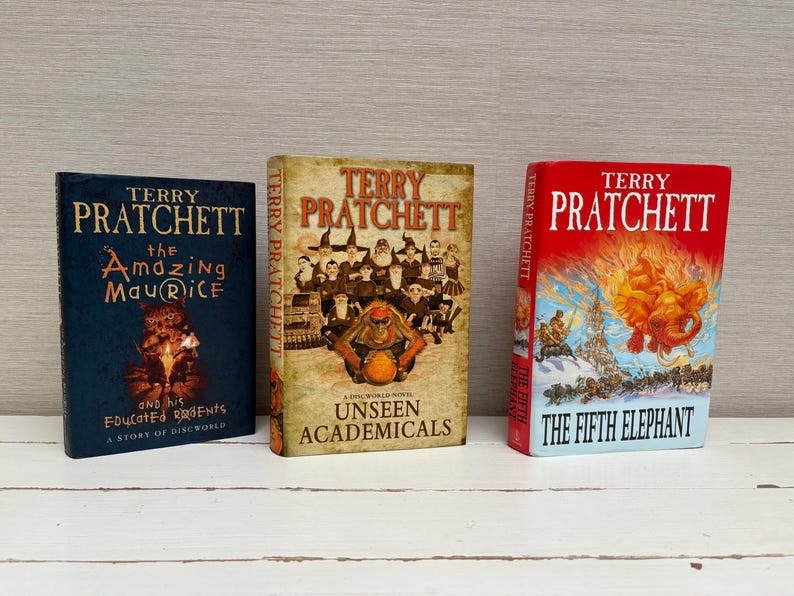 May include: Three hardcover books by Terry Pratchett, featuring colorful illustrations and titles: "The Amazing Maurice and his Educated Rodents", "Unseen Academicals", and "The Fifth Elephant".