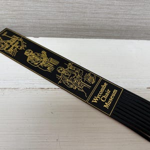 May include: Black bookmark with gold illustrations of woodworking and the text "Wycombe Chair Museum." The bookmark has a series of black fringe at the bottom. The illustrations depict chair-making processes.