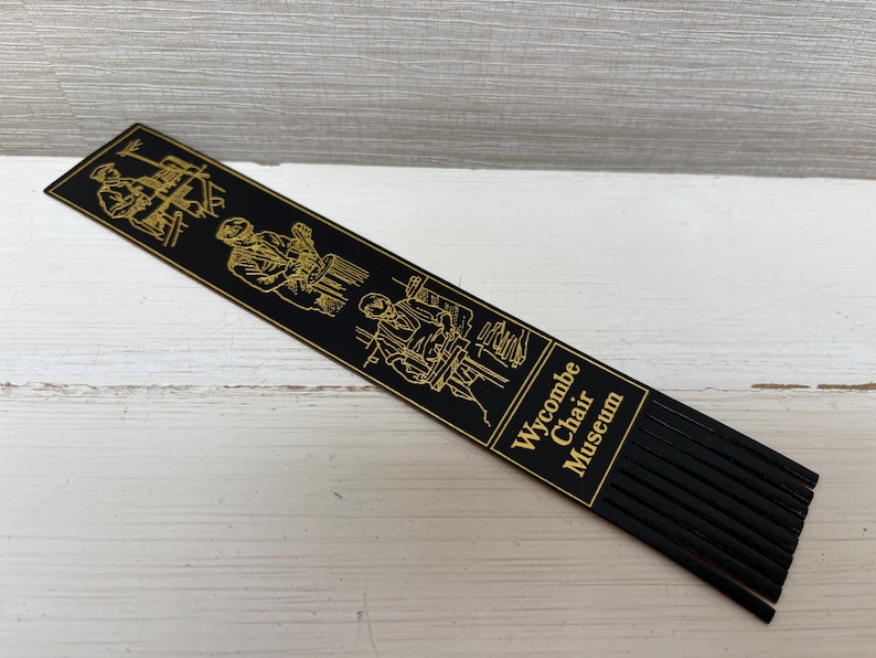 May include: Black bookmark with gold illustrations of woodworking and the text "Wycombe Chair Museum." The bookmark has a series of black fringe at the bottom. The illustrations depict chair-making processes.