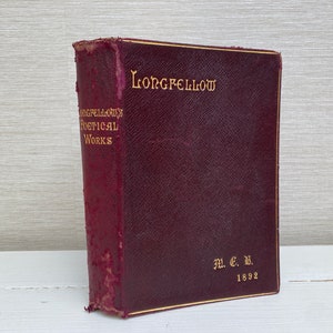 Puede incluir: Un libro encuadernado en cuero burdeos con letras doradas en el lomo. El título es "LONGFELLOW" y el lomo también dice "LONGFELLOW'S POETICAL WORKS" y "M.C.M. 1892".
