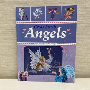 Pode incluir: Uma capa de livro com o título "Cross Stitch Angels" e um subtítulo "Mais de 30 designs inspiradores". A capa apresenta um fundo roxo com corações rosa e um design de ponto cruz de um anjo com asas.