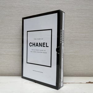 Può includere: Un libro con copertina rigida intitolato "The Story of Chanel: The Ultimate Guide to the Iconic Fashion House". Il libro ha una copertina bianca con testo nero e un bordo nero. Il dorso presenta anche il nome "Chanel".