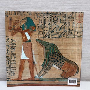 May include: An Egyptian papyrus print featuring a detailed illustration of a figure with a bird head and a jackal-headed deity. The artwork is rendered in earthy tones of brown, beige, and teal, with hieroglyphs along the borders.
