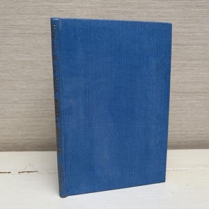 May include: A hardcover book with a blue linen cover stands upright. The spine features gold lettering. The book is positioned on a white surface against a light wood-grain background. The book's cover is a solid, medium blue.