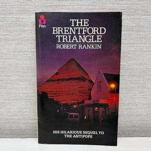 May include: A paperback copy of "The Brentford Triangle" by Robert Rankin. The cover shows a red-lit pyramid, a white house, and the text "HIS HILARIOUS SEQUEL TO THE ANTIPOPE". The book has a Pan logo in the top left corner.