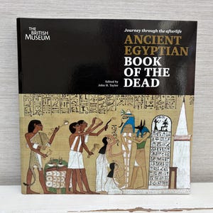 May include: A hardcover book titled "ANCIENT EGYPTIAN BOOK OF THE DEAD" from The British Museum. The cover features a black top section with the title and a lower section with an ancient Egyptian illustration in shades of tan, brown, and blue.