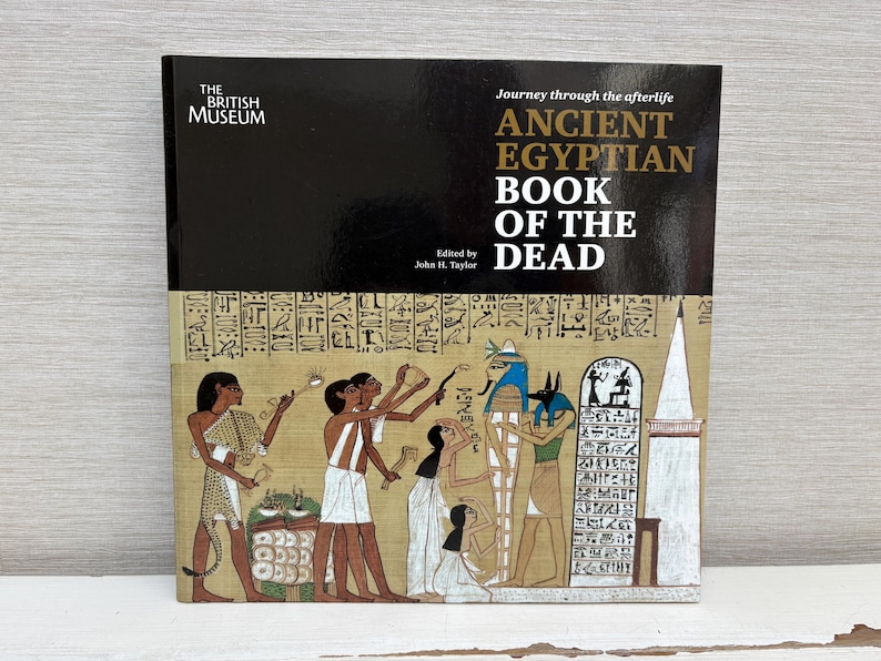 May include: A hardcover book titled "ANCIENT EGYPTIAN BOOK OF THE DEAD" from The British Museum. The cover features a black top section with the title and a lower section with an ancient Egyptian illustration in shades of tan, brown, and blue.