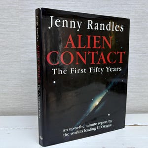 May include: A black book with the title "Alien Contact: The First Fifty Years" by Jenny Randles. The cover features a galaxy with stars and a white text overlay that reads "An up-to-the minute report by the world's leading UFOlogist."