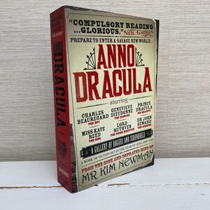 Może przedstawiać: Czerwona książka z vintage'owym plakatem z tytułem "Anno Dracula" i obsadą postaci. Książka jest autorstwa pana Kim Newman.