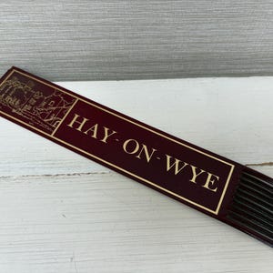 May include: A burgundy bookmark with gold lettering and a gold border. The text "HAY-ON-WYE" is displayed prominently. The top of the bookmark features a gold illustration of buildings. The bottom has black fringe.