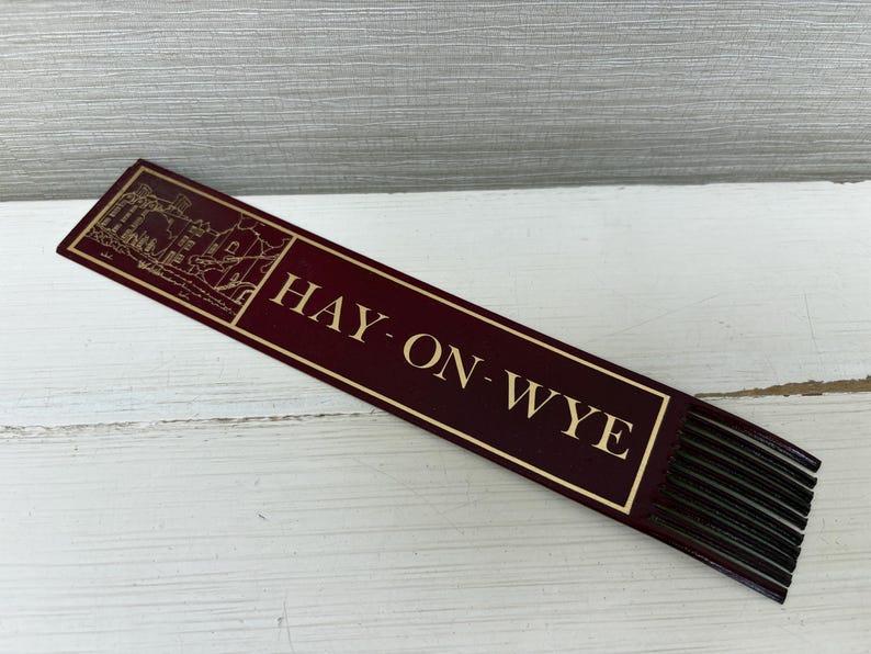 May include: A burgundy bookmark with gold lettering and a gold border. The text "HAY-ON-WYE" is displayed prominently. The top of the bookmark features a gold illustration of buildings. The bottom has black fringe.