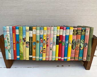 May include: A collection of vintage children's books with colorful covers, displayed on a wooden book stand. The books feature titles like "Fireside Tales" and "The Adventure Stories". The spines are a variety of colors, including red, blue, and green.