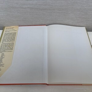 May include: An open book with a red spine and a tan cover. The left page has text in black, and the right page is blank. The book is on a white surface with a gray background.