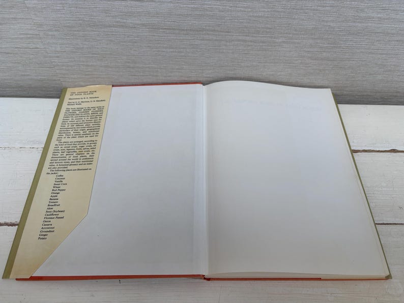 May include: An open book with a red spine and a tan cover. The left page has text in black, and the right page is blank. The book is on a white surface with a gray background.