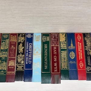 May include: A collection of colorful bookmarks with gold foil accents and fringed bottoms. Each bookmark features a different city or place name, including "London," "Ripon," and "East Yorkshire."
