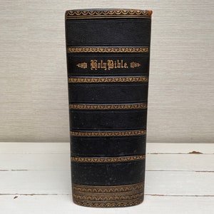 May include: A black leather-bound book with gold lettering that reads "Holy Bible". The book has a decorative pattern of gold lines and designs.