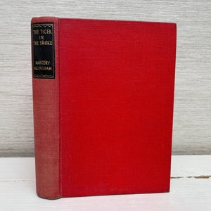 Puede incluir: Un libro de tapa dura rojo titulado "The Tiger in the Smoke" de Margery Allingham.