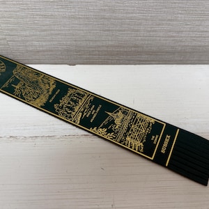 May include: A dark green bookmark with gold-colored illustrations and text. The bookmark features images of Sussex landmarks, including Beachy Head, River Arun, and Arundel Castle. The word "Sussex" is printed in gold.