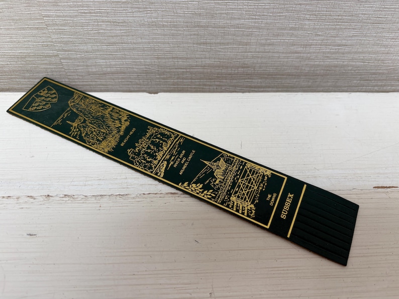 May include: A dark green bookmark with gold-colored illustrations and text. The bookmark features images of Sussex landmarks, including Beachy Head, River Arun, and Arundel Castle. The word "Sussex" is printed in gold.