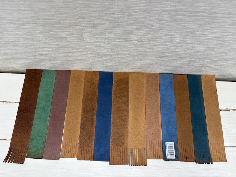 May include: A collection of leather bookmarks in various colors, including brown, green, blue, and tan. Each bookmark has a fringed bottom and a rectangular shape. Some have a textured appearance, and one has a barcode sticker.