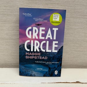 Op de afbeelding: Een paperback boek getiteld "Great Circle" van Maggie Shipstead. De omslag toont een bergketen met een vliegtuig dat erboven vliegt. Het boek heeft positieve recensies ontvangen van The Guardian, The New York Times, The Times en Curtis Sittenfeld. Het werd ook genomineerd voor de Women's Prize for Fiction in 2021.