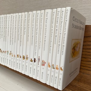 Puede incluir: Una colección de 23 libros blancos con títulos en negro, que presentan ilustraciones de personajes de Beatrix Potter. Los libros están dispuestos verticalmente en un estante de madera, con los títulos visibles. Los libros forman parte de la serie "Cecily Parsley's Nursery Rhymes".