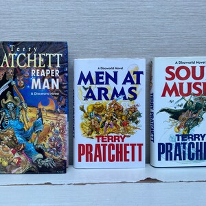 May include: Three paperback books by Terry Pratchett, all with white covers and colorful illustrations. The titles are "Reaper Man", "Men at Arms", and "Soul Music".
