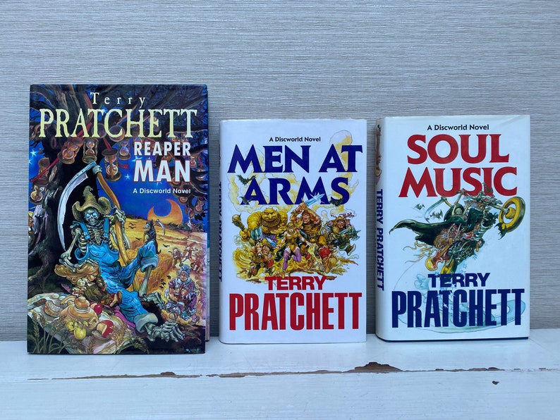 May include: Three paperback books by Terry Pratchett, all with white covers and colorful illustrations. The titles are "Reaper Man", "Men at Arms", and "Soul Music".