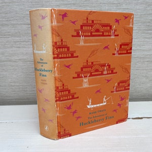 Op de afbeelding: Een vintage hardcover boek met een oranje omslag met een herhalend patroon van stoomboten en vogels. De titel "De avonturen van Huckleberry Finn" van Mark Twain staat op de rug gedrukt.