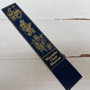 May include: A navy blue bookmark with gold illustrations of woodworking and the text "Wycombe Chair Museum". The bookmark has a fringed bottom and is lying on a white wooden surface.