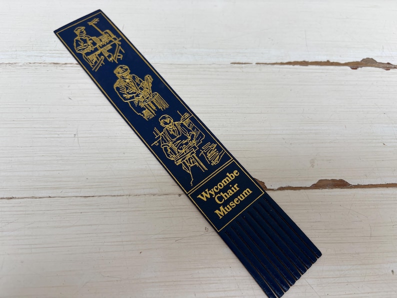 May include: A navy blue bookmark with gold illustrations of woodworking and the text "Wycombe Chair Museum". The bookmark has a fringed bottom and is lying on a white wooden surface.