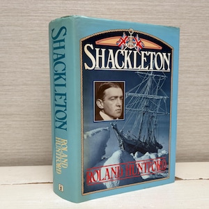 May include: A hardback book titled "Shackleton" by Roland Huntford. The cover shows a portrait, a ship illustration, and a blue and red design. The book's spine displays the title vertically in blue.