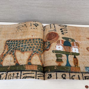 May include: Open book with ancient Egyptian artwork. The illustration features a spotted animal, possibly a cow or bull, and hieroglyphs. The artwork is in shades of brown, green, and blue, with a table of offerings.