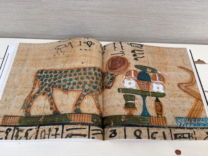 May include: Open book with ancient Egyptian artwork. The illustration features a spotted animal, possibly a cow or bull, and hieroglyphs. The artwork is in shades of brown, green, and blue, with a table of offerings.
