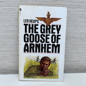 Op de afbeelding: Een paperback boekomslag met een witte achtergrond en zwarte tekst. De titel is "The Grey Goose of Arnhem" door Leo Heaps. De omslag toont een zwart-witte illustratie van een soldaat in een veld.
