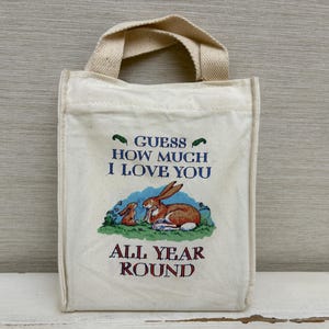 May include: Off-white canvas tote bag with a cream-colored handle. The bag features a colorful illustration of two rabbits and the text "Guess How Much I Love You All Year Round" in blue and red.