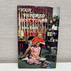 May include: A book titled "Your Hundred Best Tunes" by Alan Keith. The cover features a woman and a girl sitting in a living room with a record player and records.