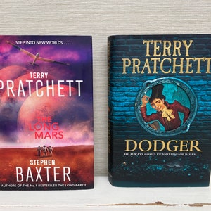 May include: Two paperback books by Terry Pratchett. The book on the left is titled "The Long Mars" and features a red planet with a small airplane flying over it. The book on the right is titled "Dodger" and features a man in a top hat and red jacket looking out of a round window.
