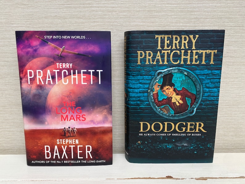 May include: Two paperback books by Terry Pratchett. The book on the left is titled "The Long Mars" and features a red planet with a small airplane flying over it. The book on the right is titled "Dodger" and features a man in a top hat and red jacket looking out of a round window.