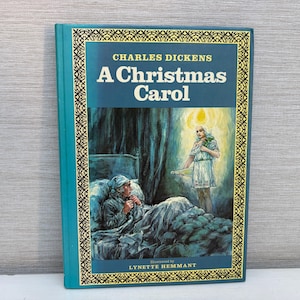 Pode incluir: Um livro de capa dura intitulado A Christmas Carol de Charles Dickens, ilustrado por Lynette Hemmant. A capa azul-petróleo apresenta uma borda dourada decorativa e uma ilustração de uma pessoa na cama visitada por uma figura brilhante.