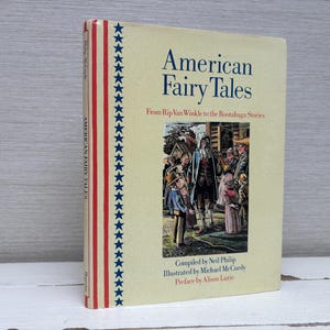 以下が含まれることがあります： 「American Fairy Tales」というタイトルのハードカバー本。クリーム色の表紙で、人々や建物のイラストと青い文字が描かれています。背表紙には赤と白のストライプと青い星があります。