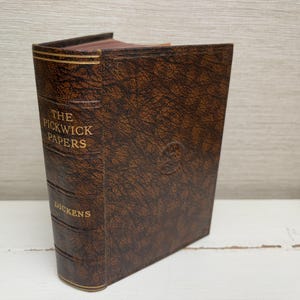 May include: A vintage edition of "The Pickwick Papers" by Dickens. The book has a brown, textured cover with gold lettering on the spine and front. The edges of the pages are visible, and the book is standing upright.