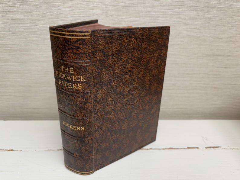 May include: A vintage edition of "The Pickwick Papers" by Dickens. The book has a brown, textured cover with gold lettering on the spine and front. The edges of the pages are visible, and the book is standing upright.