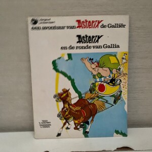 Op de afbeelding: Een vintage Asterix-stripboekomslag met een cartoonillustratie van Asterix en Obelix in een wagen getrokken door twee paarden. De omslag is wit met blauwe en gele accenten. De titel luidt "Asterix de Gallier" en "Asterix en de ronde van Gallia".