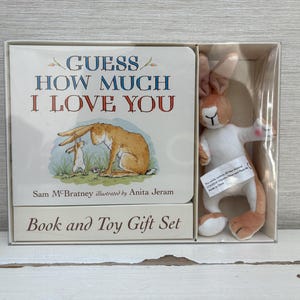 May include: A boxed "Guess How Much I Love You" book and toy gift set. The book cover features a watercolor illustration of two hares. A plush hare toy with white and tan fur is included in the box.