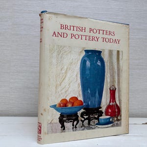 Może przedstawiać: Zabytkowa książka zatytułowana "British Potters and Pottery Today" z ilustracją na okładce przedstawiającą niebieski wazon, czerwony wazon i miskę pomarańczy. Książka jest umieszczona na teksturowanym tle.
