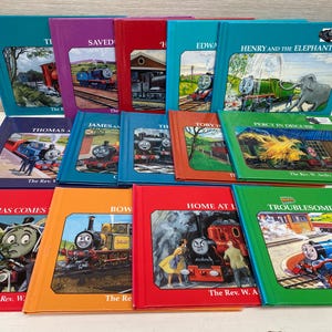 May include: A collection of ten colorful children's books featuring Thomas the Tank Engine. The books are arranged in two rows, with the titles visible on the spines. The titles include "Thomas Comes", "Bow", "Home at Last", "Troublesome Engines", "Percy in Disguise", "Toby", "James and the Coaches", "Thomas and the Breakdown Train", "Saved from Scrap", and "Henry and the Elephant".